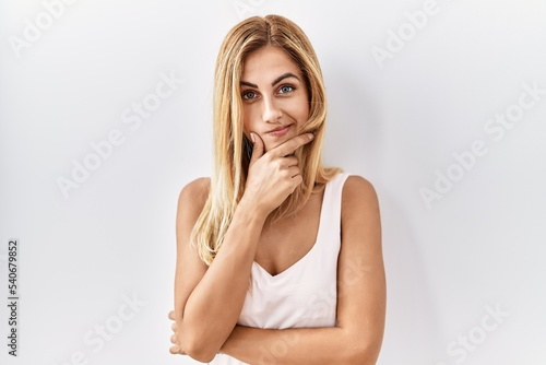 Blonde beautiful young woman standing over white isolated background looking confident at the camera with smile with crossed arms and hand raised on chin. thinking positive.