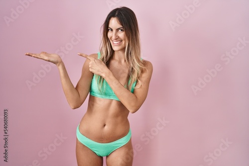 Young hispanic woman wearing bikini over pink background amazed and smiling to the camera while presenting with hand and pointing with finger. © Krakenimages.com