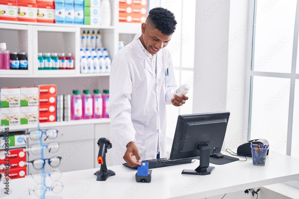 Young latin man pharmacist using credit card and data phone holding pills bottle at pharmacy Young latin man pharmacist using credit card and data phone holding pills bottle at pharmacy