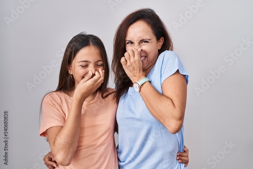 Young mother and daughter standing over white background smelling something stinky and disgusting, intolerable smell, holding breath with fingers on nose. bad smell © Krakenimages.com