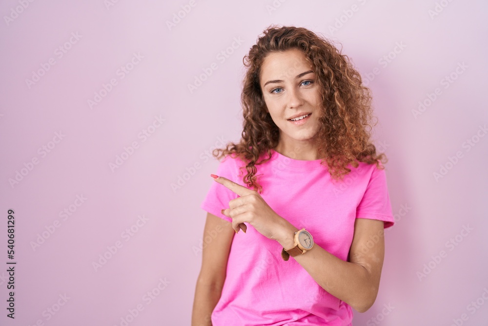 Young caucasian woman standing over pink background cheerful with a smile on face pointing with hand and finger up to the side with happy and natural expression Young caucasian woman standing over pink background cheerful with a smile on face pointing with hand and finger up to the side with happy and natural expression