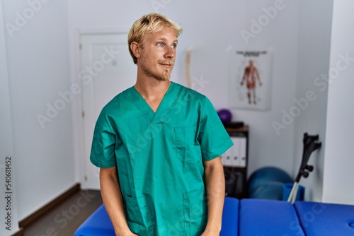 Young blond man wearing physiotherapist uniform standing at clinic looking away to side with smile on face, natural expression. laughing confident.