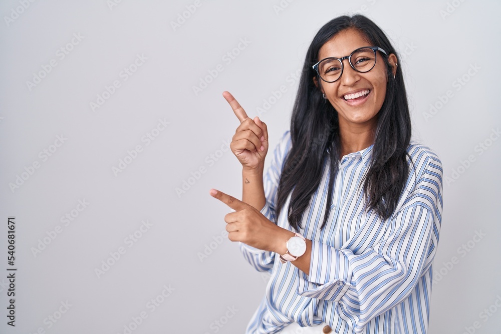 Young hispanic woman wearing glasses smiling and looking at the camera pointing with two hands and fingers to the side.
