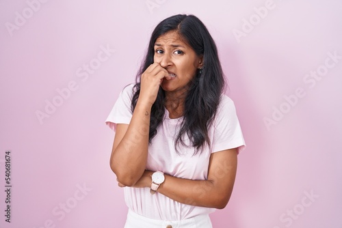 Young hispanic woman standing over pink background looking stressed and nervous with hands on mouth biting nails. anxiety problem.