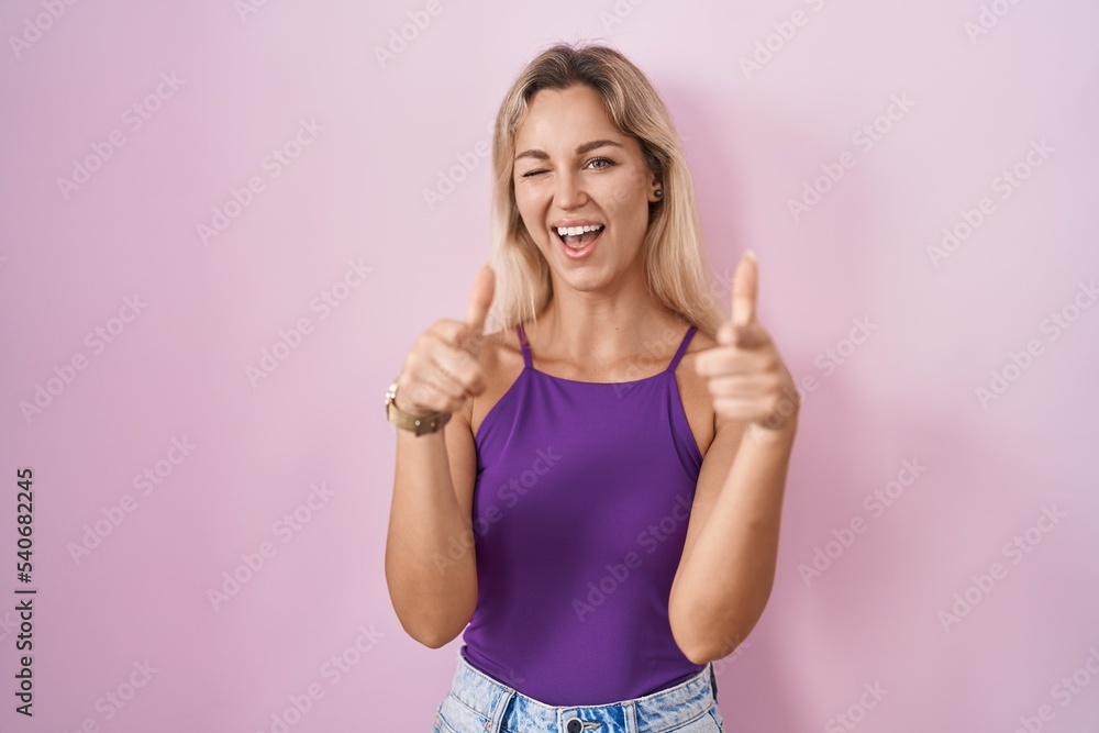 Young blonde woman standing over pink background pointing fingers to camera with happy and funny face. good energy and vibes.