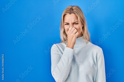 Young caucasian woman standing over blue background smelling something stinky and disgusting, intolerable smell, holding breath with fingers on nose. bad smell photo