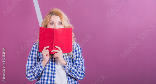 Young girl blonde stands background of pink wall, covered half her face with book and looks with large surprised eyes at camera.