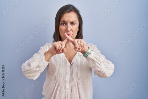Middle age hispanic woman standing over blue background rejection expression crossing fingers doing negative sign