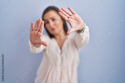 Middle age hispanic woman standing over blue background doing frame using hands palms and fingers, camera perspective