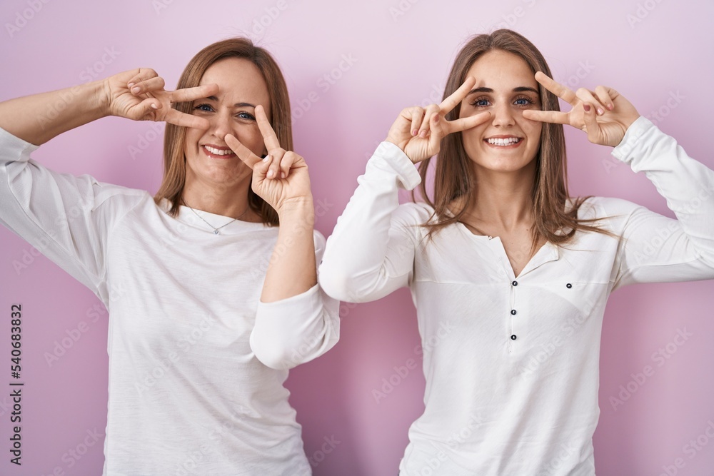 Middle age mother and young daughter standing over pink background doing peace symbol with fingers over face, smiling cheerful showing victory Middle age mother and young daughter standing over pink background doing peace symbol with fingers over face, smiling cheerful showing victory