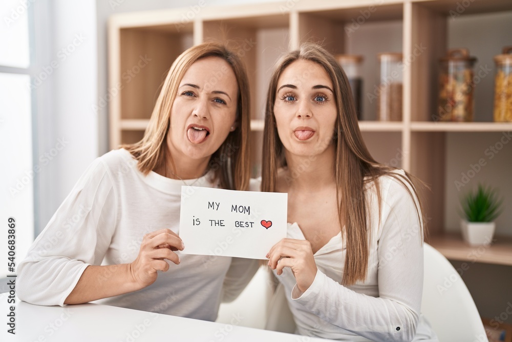 Foto de Middle age mother and young daughter holding my mom is the best banner sticking tongue