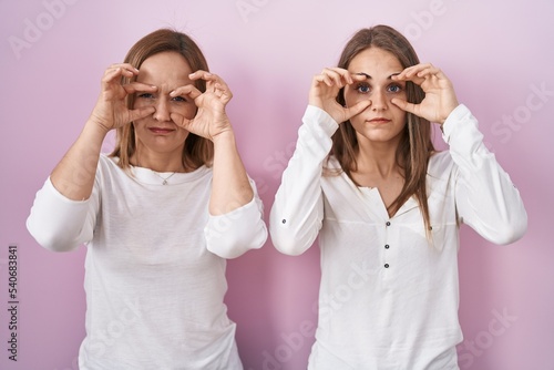 Middle age mother and young daughter standing over pink background trying to open eyes with fingers, sleepy and tired for morning fatigue