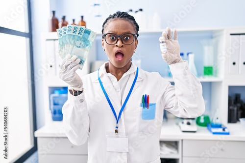 African woman with braids working at scientist laboratory holding money in shock face  looking skeptical and sarcastic  surprised with open mouth