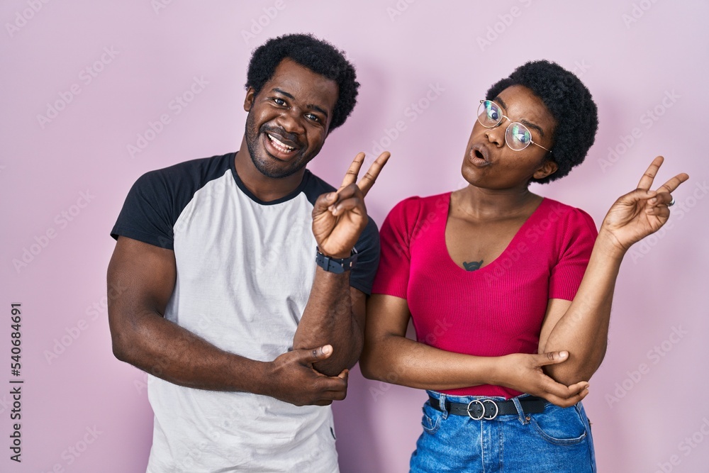 Young african american couple standing over pink background smiling with happy face winking at the camera doing victory sign with fingers. number two. Young african american couple standing over pink background smiling with happy face winking at the camera doing victory sign with fingers. number two.