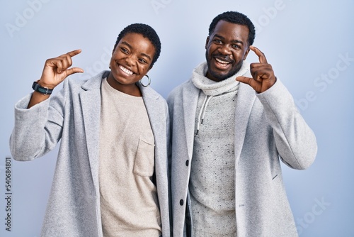 Young african american couple standing over blue background together smiling and confident gesturing with hand doing small size sign with fingers looking and the camera. measure concept.