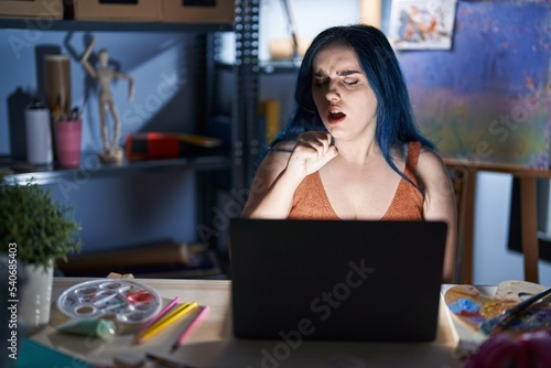 Young modern girl with blue hair sitting at art studio with laptop at night feeling unwell and coughing as symptom for cold or bronchitis. health care concept.