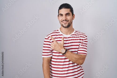 Hispanic man with long hair standing over isolated background cheerful with a smile on face pointing with hand and finger up to the side with happy and natural expression