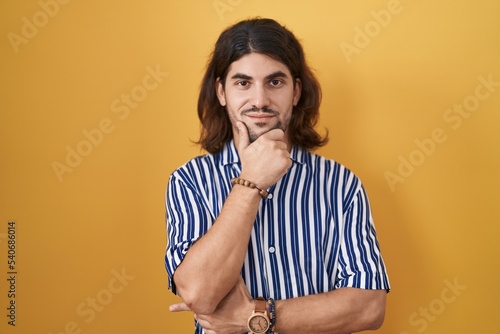 Hispanic man with long hair standing over yellow background looking confident at the camera with smile with crossed arms and hand raised on chin. thinking positive.