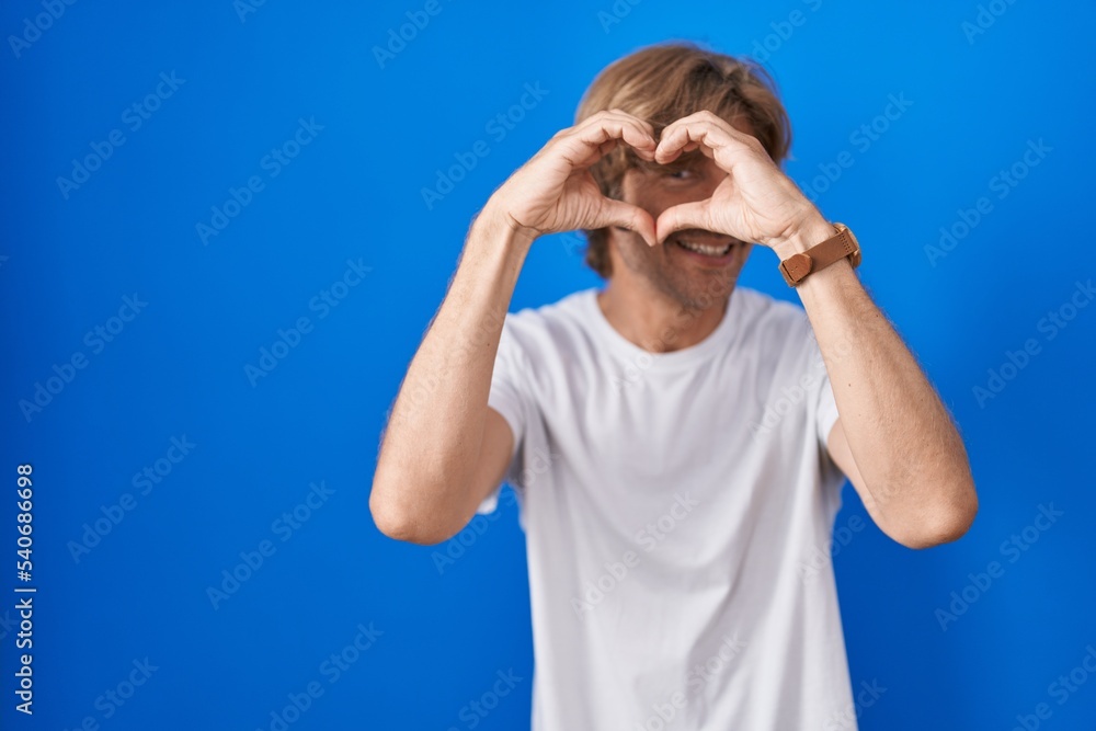 Middle age man standing over blue background doing heart shape with hand and fingers smiling looking through sign Middle age man standing over blue background doing heart shape with hand and fingers smiling looking through sign
