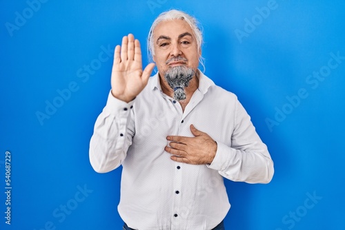 Middle age man with grey hair standing over blue background swearing with hand on chest and open palm, making a loyalty promise oath © Krakenimages.com