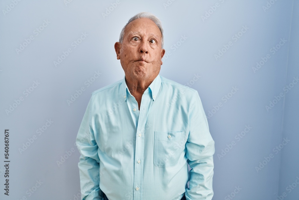 Obraz premium Senior man with grey hair standing over blue background making fish face with lips, crazy and comical gesture. funny expression.