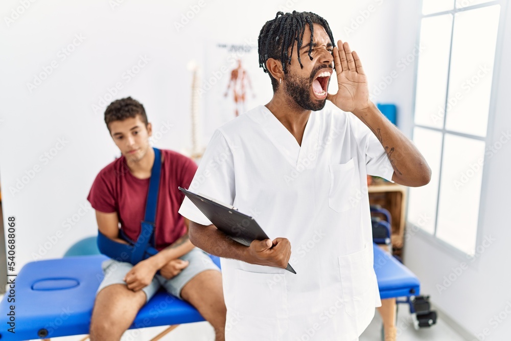Obraz premium Young hispanic man working at pain recovery clinic with a man with broken arm shouting and screaming loud to side with hand on mouth. communication concept.