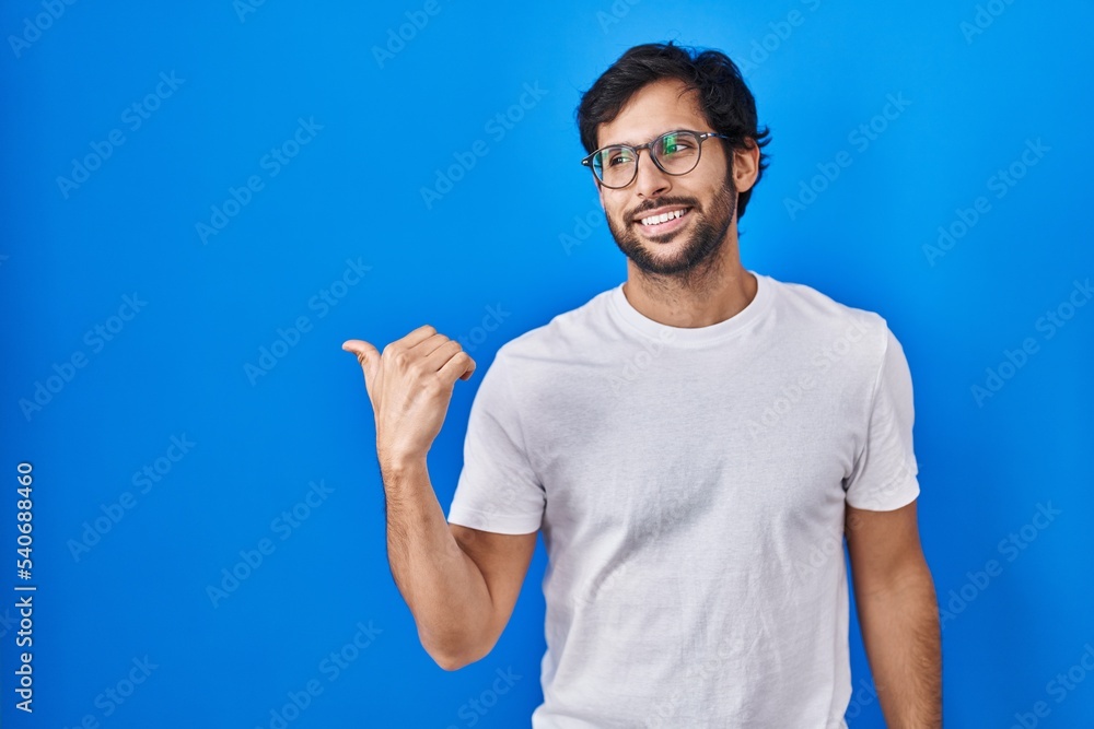Handsome latin man standing over blue background smiling with happy face looking and pointing to the side with thumb up. Handsome latin man standing over blue background smiling with happy face looking and pointing to the side with thumb up.