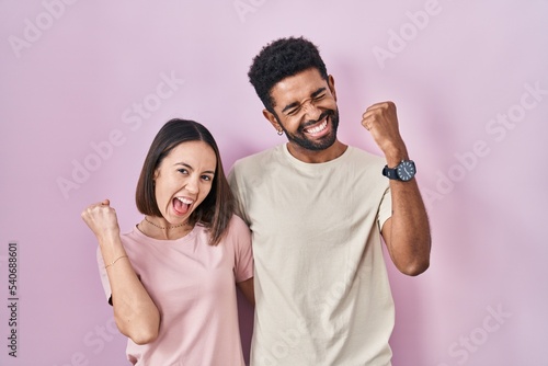 Young hispanic couple together over pink background very happy and excited doing winner gesture with arms raised, smiling and screaming for success. celebration concept.
