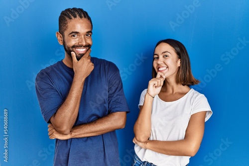 Young hispanic couple standing together looking confident at the camera smiling with crossed arms and hand raised on chin. thinking positive.