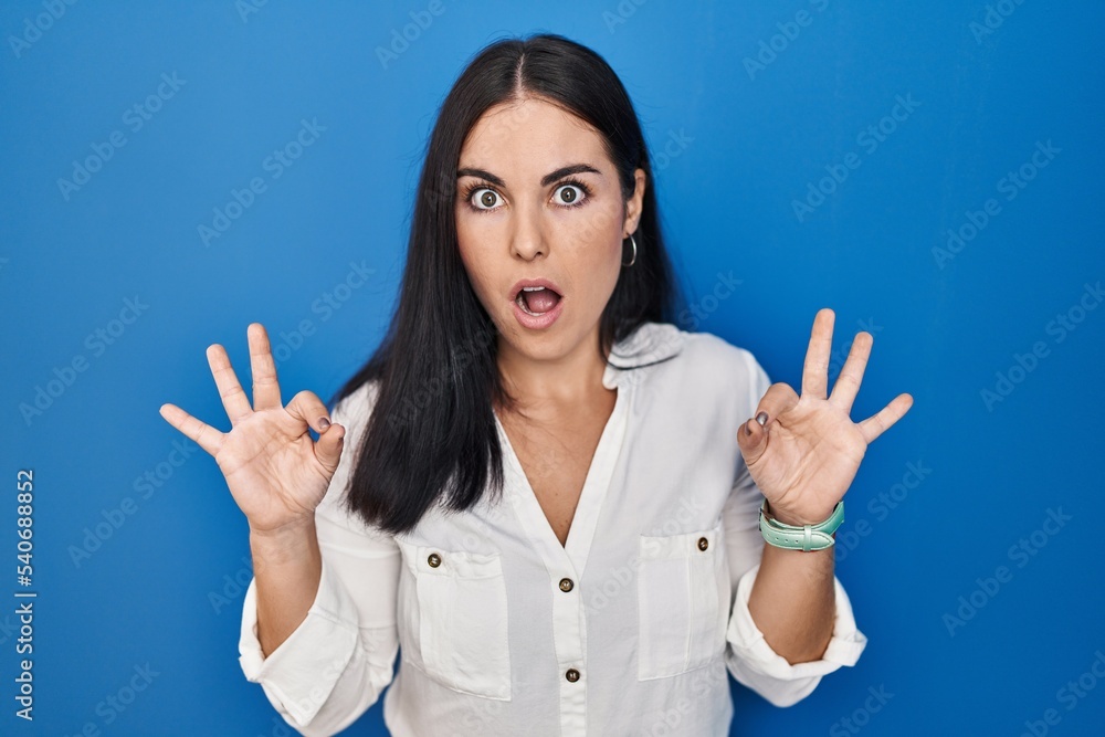 Young hispanic woman standing over blue background looking surprised and shocked doing ok approval symbol with fingers. crazy expression Young hispanic woman standing over blue background looking surprised and shocked doing ok approval symbol with fingers. crazy expression