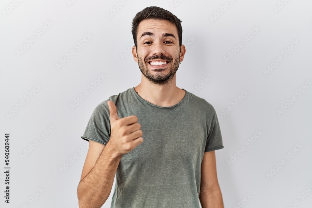 Fototapeta premium Young hispanic man with beard wearing casual t shirt over white background doing happy thumbs up gesture with hand. approving expression looking at the camera showing success.