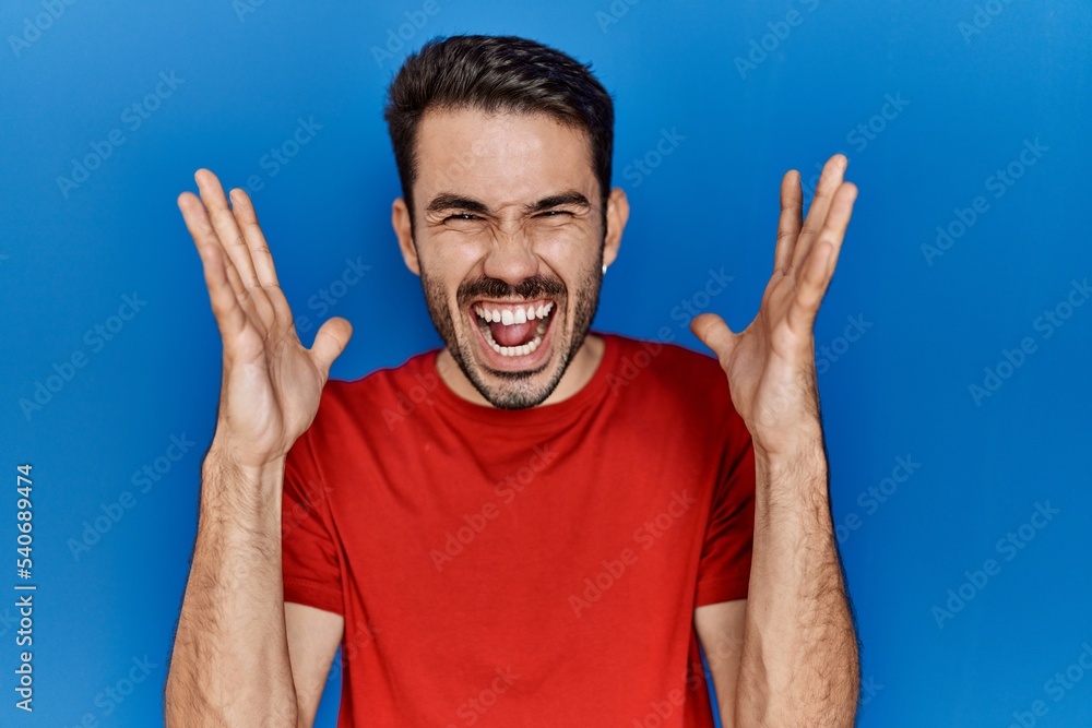 Naklejka premium Young hispanic man with beard wearing red t shirt over blue background celebrating mad and crazy for success with arms raised and closed eyes screaming excited. winner concept