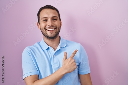 Hispanic man standing over pink background cheerful with a smile on face pointing with hand and finger up to the side with happy and natural expression