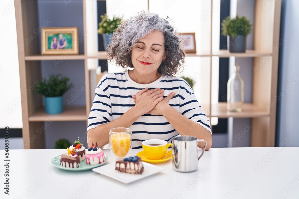 Middle age woman with grey hair eating pastries and drinking coffee for breakfast smiling with hands on chest with closed eyes and grateful gesture on face. health concept. Middle age woman with grey hair eating pastries and drinking coffee for breakfast smiling with hands on chest with closed eyes and grateful gesture on face. health concept.