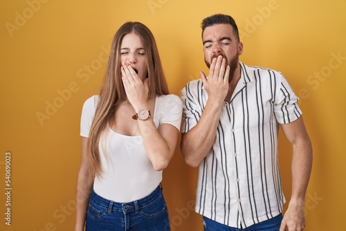 Young couple standing over yellow background bored yawning tired covering mouth with hand. restless and sleepiness.
