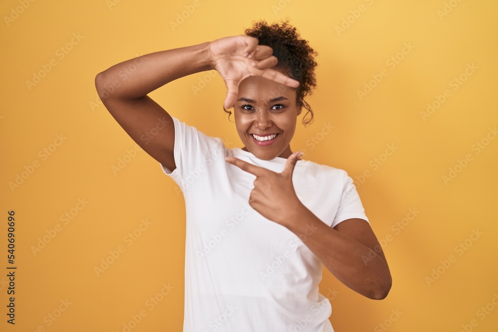 Young hispanic woman with curly hair standing over yellow background smiling making frame with hands and fingers with happy face. creativity and photography concept. Young hispanic woman with curly hair standing over yellow background smiling making frame with hands and fingers with happy face. creativity and photography concept.