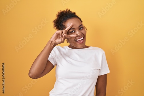Young hispanic woman with curly hair standing over yellow background doing peace symbol with fingers over face  smiling cheerful showing victory