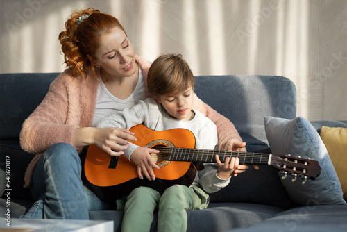 Happy mother teaching her son how to play the guitar in their living room on sunny day