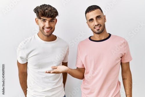 Young gay couple standing over isolated background smiling cheerful presenting and pointing with palm of hand looking at the camera.