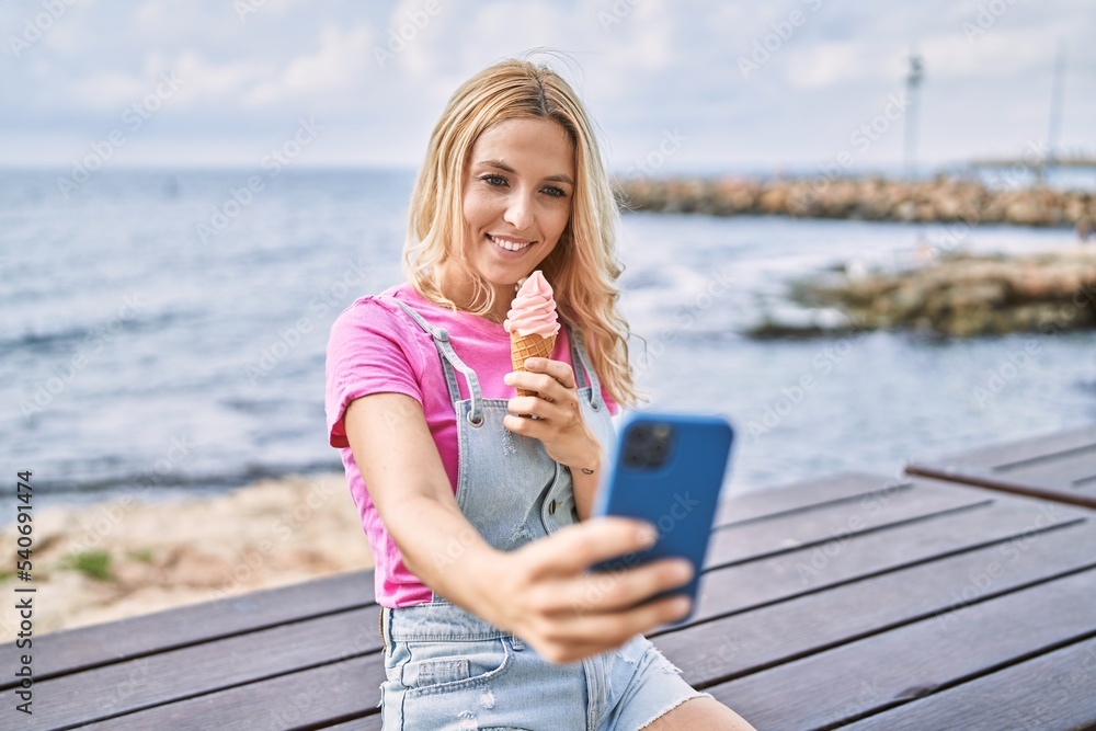 Young blonde woman eating ice cream make selfie by the smartphone at seaside
