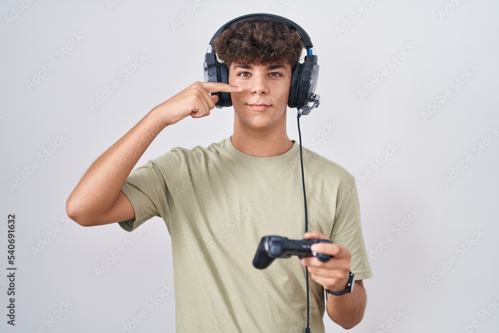Hispanic teenager playing video game holding controller pointing with hand finger to face and nose, smiling cheerful. beauty concept Hispanic teenager playing video game holding controller pointing with hand finger to face and nose, smiling cheerful. beauty concept