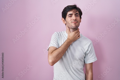 Young hispanic man standing over pink background touching painful neck, sore throat for flu, clod and infection