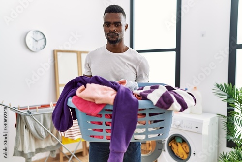 Young african man holding laundry basket relaxed with serious expression on face. simple and natural looking at the camera.