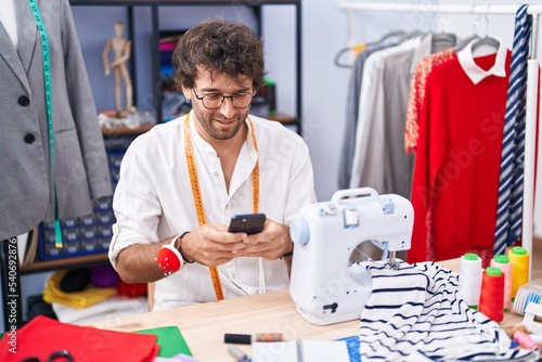 Young hispanic man tailor smiling confident using smartphone at clothing factory
