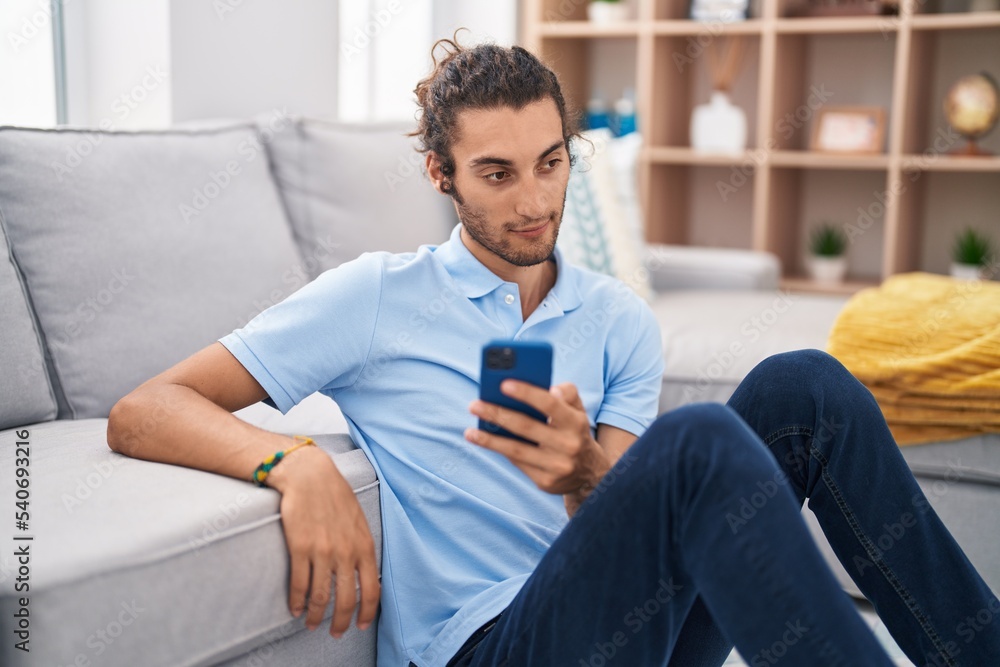 Obraz premium Young hispanic man using smartphone sitting on floor at home