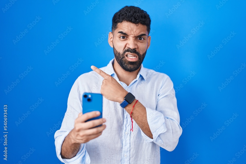 Hispanic man with beard using smartphone typing message pointing aside worried and nervous with forefinger, concerned and surprised expression Hispanic man with beard using smartphone typing message pointing aside worried and nervous with forefinger, concerned and surprised expression