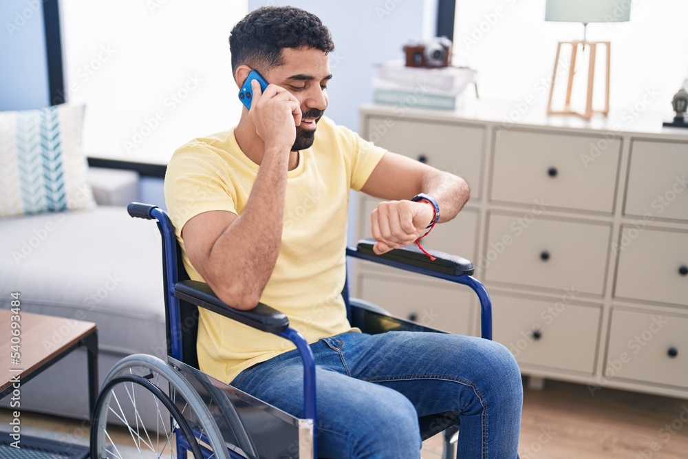 Young arab man talking on smartphone looking watch sitting on wheelchair at home