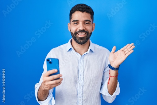 Hispanic man with beard using smartphone typing message smiling cheerful presenting and pointing with palm of hand looking at the camera.