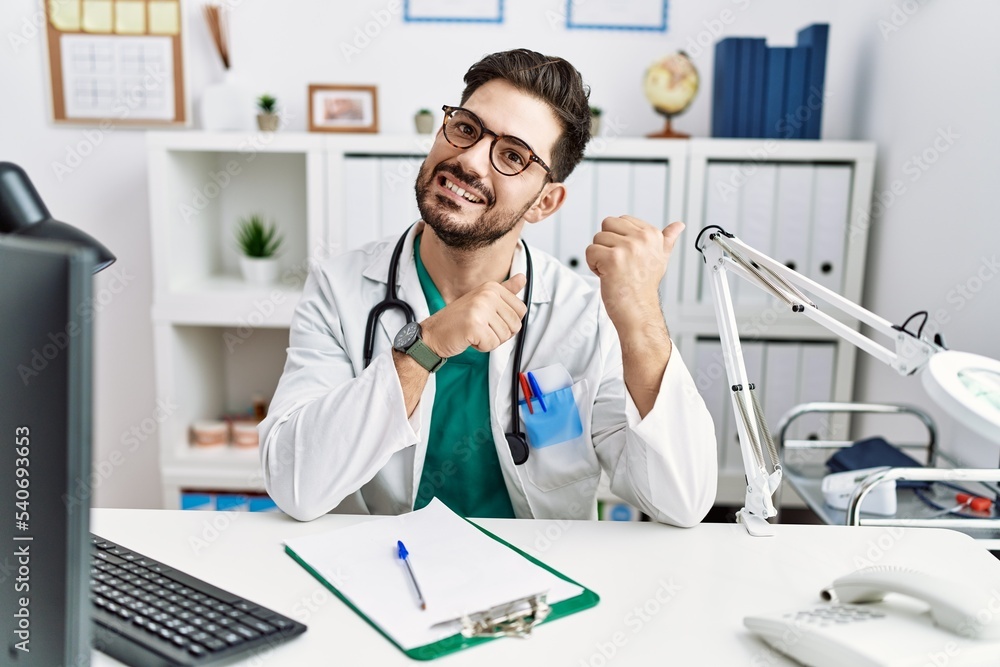 Young man with beard wearing doctor uniform and stethoscope at the clinic pointing to the back behind with hand and thumbs up, smiling confident Young man with beard wearing doctor uniform and stethoscope at the clinic pointing to the back behind with hand and thumbs up, smiling confident