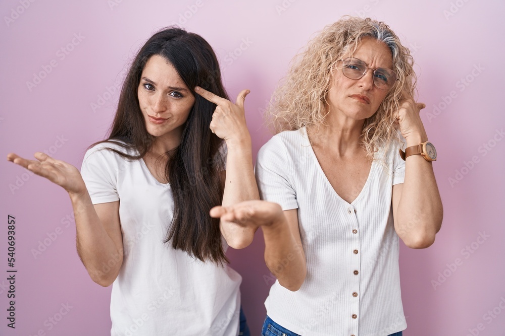 Mother and daughter standing together over pink background confused and annoyed with open palm showing copy space and pointing finger to forehead. think about it. Mother and daughter standing together over pink background confused and annoyed with open palm showing copy space and pointing finger to forehead. think about it.